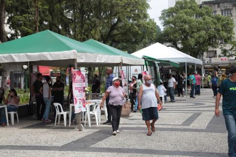 Pessoas circulam na praça com várias tendas montadas #paratodosverem