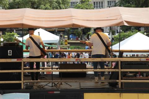 artistas (de costas) tocam no palco e pessoas e praça a frente deles #paratodosverem