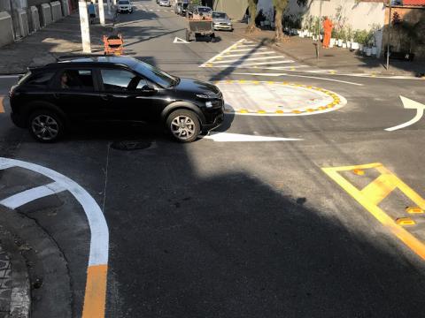 Veículo preto circula pela minirrotatória. #pracegover