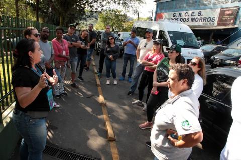 grupo de pessoas em pé em uma praça. Uma mulher, à esquerda e usando crachá fala ao grupo. #paratodosverem