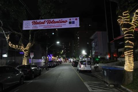 rua com árvores decoradas por cordões luminosos nos troncos. No alto e no meio da via há uma faixa onde se lê Natal Tolentino. #paratodosverem