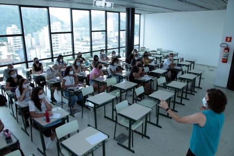 vista geral de sala de aula, com prédios e morro ao fundo. Alunos estão sentados e professora, à frente, em pé. #paratodosverem