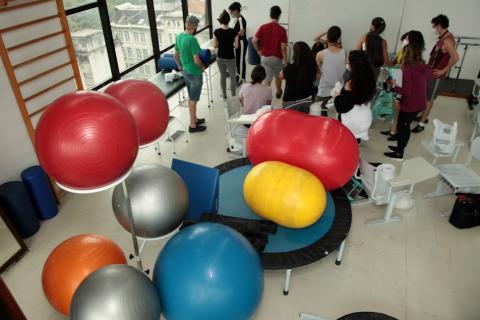 bolas de exerc´cios e outros equipamentos de fisioterapia em sala com alunos em pé. #paratodosverem