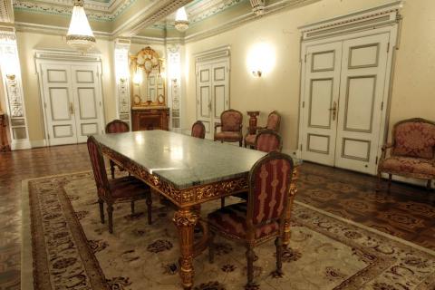 vista geral do salão nobre com mesa, cadeira, lustre, luminária, portas, tapete. #paratodosverem