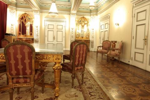 Mesa com cadeiras em primeiro plano, tapete, o piso, portas, luminária e lustres do salão nobre. #paratodosverem