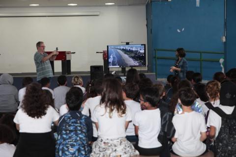 crianças estão sentadas assistindo a vídeo que passa em monitor ao fundo. Compositor está em pé ao fundo. #paratodosverem