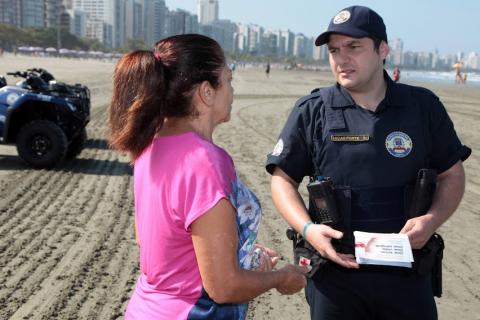 guarda segura folhetos e fala com uma mulher que está de costas para a foto. Eles estão na faixa de areia.  Atrás dele está um quadriciclo da guarda. #paratodosverem