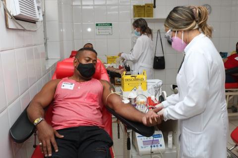 homem está reclilnado em poltrona enquanto mulher mexe em seu braço esquerdo para começar a doação. #paratodosverem