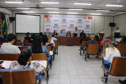 auditório com várias pessoas sentadas e algumas que se apresentam, ao fundo, falando para o público. #paratodosverem