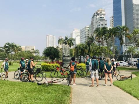 Ciclistas observam um monumento na orla da praia. #paratodosverem