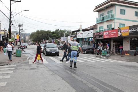 avenida com agente da cet ao centro, diante de faixas de pedestres. pessoas atravessando a rua. Estabelecimentos comerciais abertos. #paratodosverem