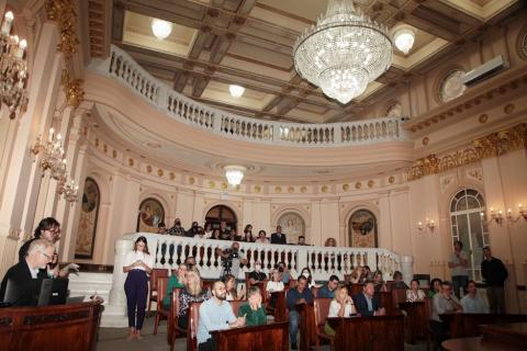 sala princesa isabel, com pessoas sentadas no plenário e auditório. Em primeiro plano, acima, um grande lustre aceso. Foto dá foco na arquitetura da sala, em curva. #paratodosverem