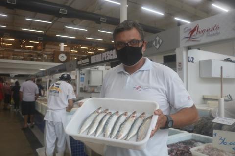 homem segura caixa de plástico com oito sardihas. Ele está à frente de um box no mercado. #paratodosverem