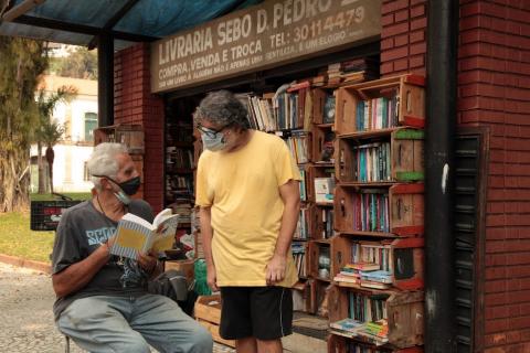 pessoa sentada lê livro ao lado de outra em pé. Ambos estão diante de uma banca de sebo. #paratodosverem