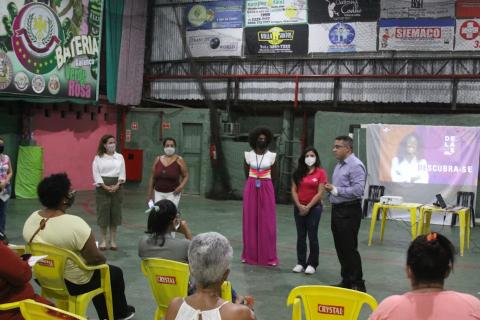 Homem fala, ao lado de quatro mulheres em pé, para o público feminino sentado. #pratodosverem