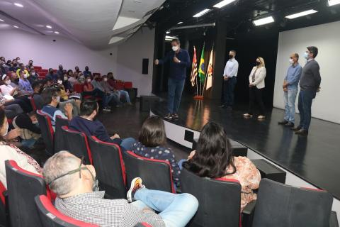 prefeiito está no palco à frente de mais pessoas. Há gente na plateia ouvindo. #paratodosverem