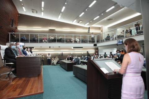 solenidade na câmara. Em primeiro plano, à direita, mulher está lendo diante de uma tribuna. Público ao fundo olha para ela. #paratodosverem