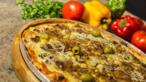 Pizza em primeiro plano com legumes ao fundo. #pracegover