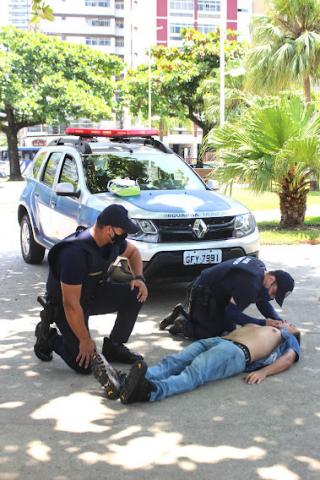 um guarda aplica o desfibrilador enquanto outro está ao lado, de apoio. Viatura da corporação está parada atrás. #paratodosverem