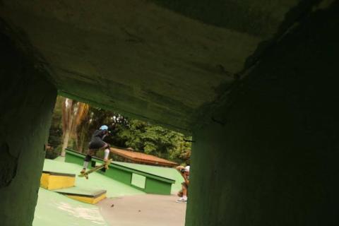 menino faz manobra de skate em degrau. Foto foi feita por baixo de uma estrutura, formando uma espécie de túnel quadrado com o garoto ao fundo. #paratodosverem