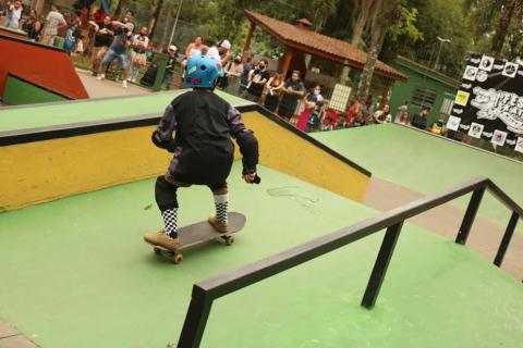 menino está com os dois pés sobre o skate indo em sentido a uma rampa. Em primeiro plano, um corrimão para manobras. #paratodosverem