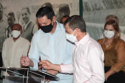O prefeito Rogério Santos e o vice-presidente do clube, José Carlos de Oliveira assinam o documento sobre uma tribuna. #paratodosverem