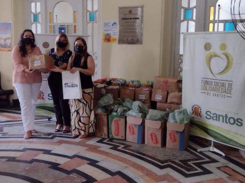 Três mulheres ao lado de caixas com doações e folder do Fundo Social #paratodosverem
