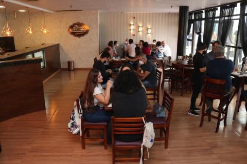 área interna da cafeteria com mesas e cadeiras. Pessoas estão sentadas e conversando. Do lado direito há um balcão, no centro as mesas, e à direita, grande janelas. #paratodosverem