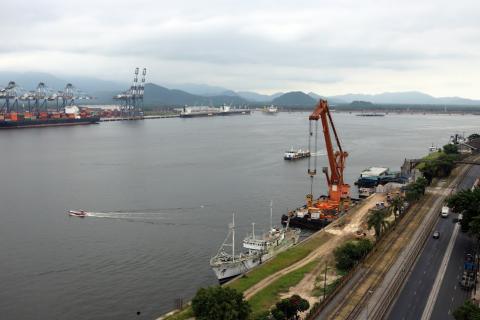 Vista de outro lado do porto, com a marguem esquerda e avenida perimetral em primeiro plano e a margem direita (Guarujá), ao fundo.Há pequenas barcas atravessando o mar e um guindaste em operação. #paratodosverem