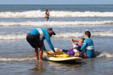 alunos é colocado sobre a prancha na beira da água com auxílio de duas pessoas. #paratodosverem