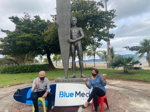 alunos da escola de surfe posam para foto em junto a uma prancha, na frente de estátua em homenagem a surfista. #paratodosverem