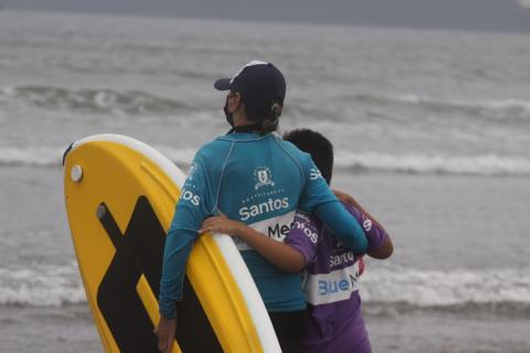 Homem segura prancha abraçado a menino. Ambos estão de frente para o mar, de costas para a foto. #paratodosverem