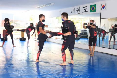 atletas treinam sobre tatame. em primeiro plano, Nathan treina dando chutes nos braços protegidos de colega. #paratodosverem