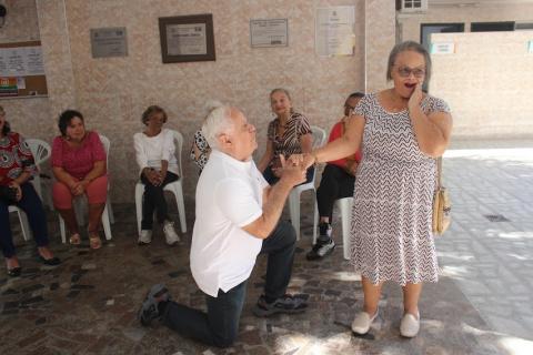 casal encenando. O homem está ajoelhado segurando a mão direita da mulher. Ela está com a outra mão na bochecha, como se estivesse surpresa. #paratodosverem