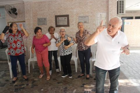 homem está em pé em primeiro plano com o dedo apontado para cima. Atrás dele, cinco mulheres espantadas com seu movimento. #paratodosverem