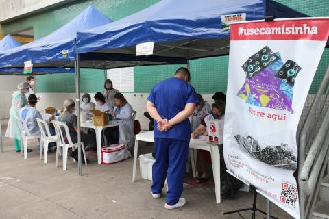 tendas montadas em frente a posto de saúde com pessoas sentadas e em pé. À direita, um banner com a #usecamisinha. #paratodosverem