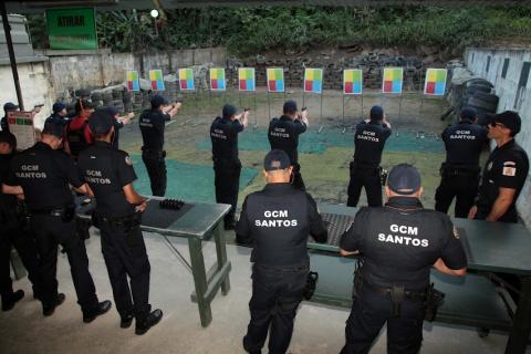 guardas, todos de costas, estão em pé, alguns diantes de mesas. Outros, com as armas na mão em posição de tiro, mirando alvos ao fundo. #paratodosverem