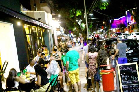 Pessoas circulando pela rua durante a noite. Há também público sentado em mesas de bar na calçada. #paratodosverem