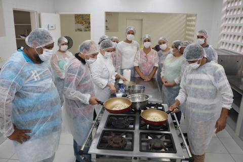 grupo de alunos está em volta de um fogão industrial. Três pessoas estão passando frigideiras no fogo. #paratodosverem