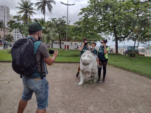 surfistas param diante de monumento de leão no jardim da orla. Um homem, de costa, fotografa três pessoas no leão. #paratodosverem