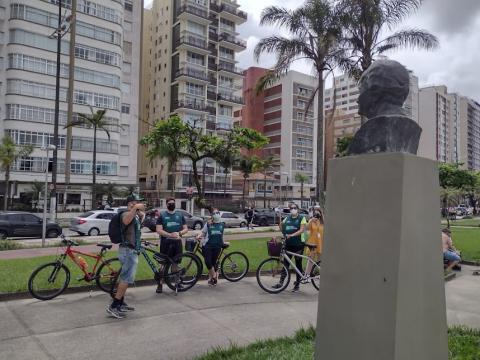 ciclistas observam busto que aparece de costas para foto em primeiro plano. Ao fundo, prédios da orla. #paratodosverem