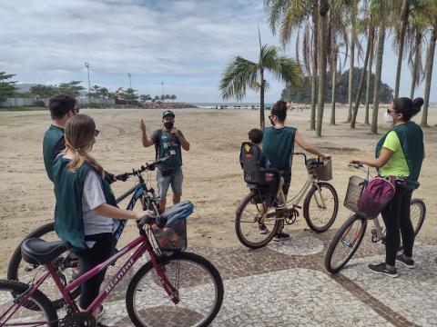 /ciclistas estão entre a calçada e a faixa de areia. Eles seguram as bikes, desmontados. Um homem, na areia fala ao grupo. Ao fundo, palmeiras da praia do Gonzaga. #paratodosverem