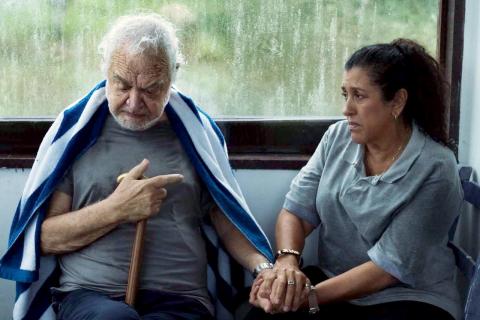 cena de três verões, com uma mulher segurando a mão de um idoso que segura uma bengala. Ele olha para baixo. Os dois estão sentados. #paratodosverem