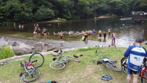 área verde com bicicletas no chão e pessoas tomando banho na cachoeira ao fundo. #paratodosverem