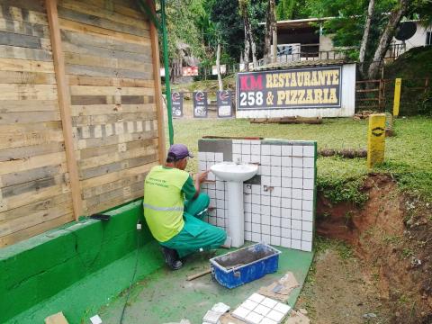 funcionário está mexendo em mureta próxima a uma pia. Ao fundo há uma placa indicando um restaurante. #paratodosverem