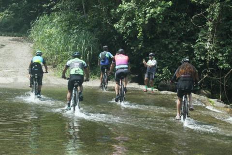 ciclistas de costas para a goto passam sobre trecho da trilha com riacho. #paratodosverem