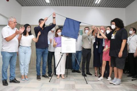 várias pessoas reunidas em torno de uma placa. Duas pessoas puxam o pano que cobre essa placa, num ato de inauguração. #paratodosverem