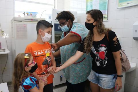 mãe está com dois filhos na sala de vacinação. Enquanto uma mulher aplica a dose no braço do garoto, a menina mais nova segura a mão dele. #paratodosverem