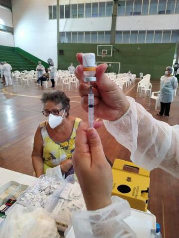 Seringa em primeiríssimo plano, com idosa sentada ao fundo. #paratodosverem