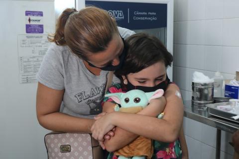 mãe ampara com abraço menina que acaba de tomar vacina. A menina também abraça um boneco. #paratodosverem